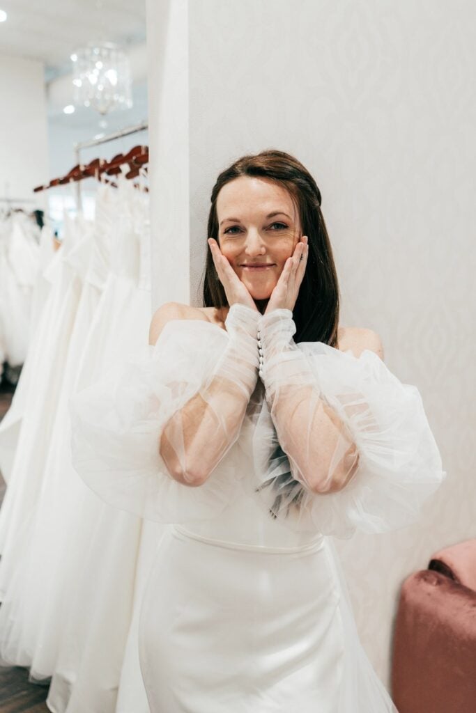 Bride smiling in a white wedding dress with sheer puff sleeves during her bridal fitting
